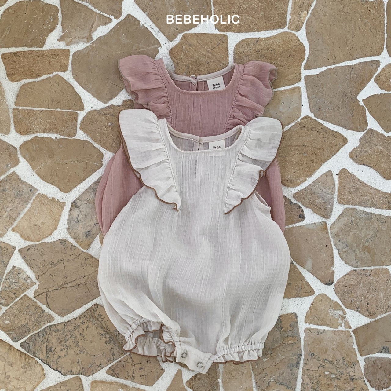 Zwei Baby-Outfits auf einem strukturierten Mosaikboden: ein rosa Oberteil mit Rüschen und ein beiger Bebe Holic Lily Wing Body auf der linken Seite und ein weißer Strampler mit Rüschen auf der rechten Seite.