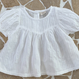 Eine weiße, kurzärmelige Bebe Holic Raoul Babybluse mit feinen Querstreifen und luftige Ärmelgestaltung, flach auf einem beige-braunen unregelmäßigen Fliesenboden liegend.