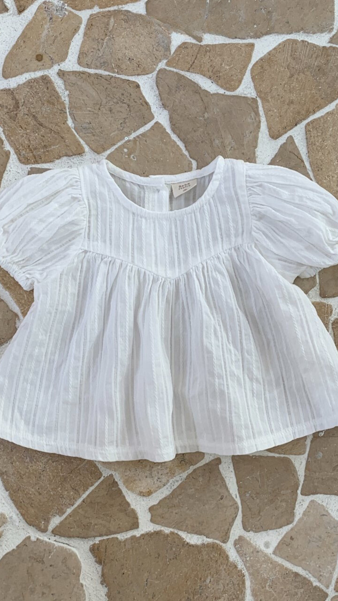 Eine weiße, kurzärmelige Bebe Holic Raoul Babybluse mit feinen Querstreifen und luftige Ärmelgestaltung, flach auf einem beige-braunen unregelmäßigen Fliesenboden liegend.