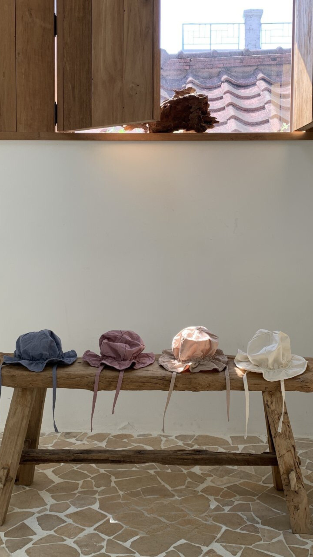 Eine Holzbank steht an einer cremefarbenen Wand und unter einem Fenster mit Holzläden. Vier Crunch Bucket Hats von Bebe Holic in Blau, Rosa, Beige und Weiß sind ordentlich auf der Bank angeordnet. Der Boden besteht aus unregelmäßig geformten Steinfliesen, die die natürliche Farbpalette des Raumes hervorheben.
