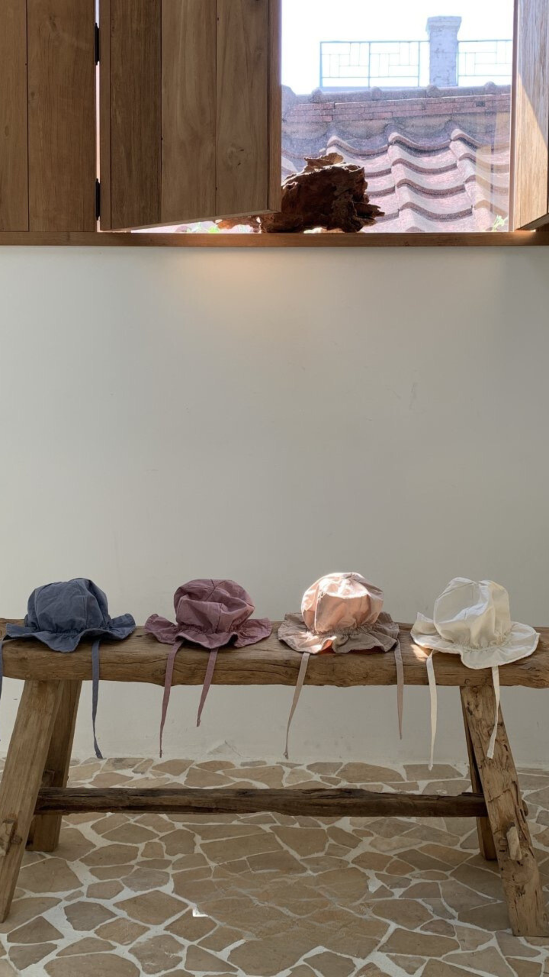 Eine rustikale Holzbank steht an einer hellen Wand unter einem Fenster mit Holzläden. Vier Crunch Bucket Hats in Blau, Lila, Rosa und Weiß von Bebe Holic sind ordentlich auf der Bank platziert. Sonnenlicht strömt durch das Fenster herein, wirft einen warmen Schein auf die Szene und verstärkt die natürliche Farbpalette.