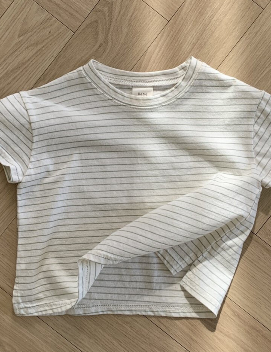 Ein weißes, kurzärmliges, kurzes T-Shirt mit dünnen schwarzen Querstreifen liegt ordentlich auf einem hölzernen Boden mit Fischgrätenmuster. Der Saum dieses Bebe Holic Basic ST Tee ist leicht angehoben und zeigt ein wenig den Stoff auf der Innenseite. Die Beleuchtung erzeugt sanfte Schatten auf dem Boden und unterstreicht seinen klassischen Stil.