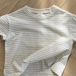 Ein weißes, kurzärmliges, kurzes T-Shirt mit dünnen schwarzen Querstreifen liegt ordentlich auf einem hölzernen Boden mit Fischgrätenmuster. Der Saum dieses Bebe Holic Basic ST Tee ist leicht angehoben und zeigt ein wenig den Stoff auf der Innenseite. Die Beleuchtung erzeugt sanfte Schatten auf dem Boden und unterstreicht seinen klassischen Stil.