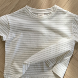 Ein weißes, kurzärmliges, kurzes T-Shirt mit dünnen schwarzen Querstreifen liegt ordentlich auf einem hölzernen Boden mit Fischgrätenmuster. Der Saum dieses Bebe Holic Basic ST Tee ist leicht angehoben und zeigt ein wenig den Stoff auf der Innenseite. Die Beleuchtung erzeugt sanfte Schatten auf dem Boden und unterstreicht seinen klassischen Stil.