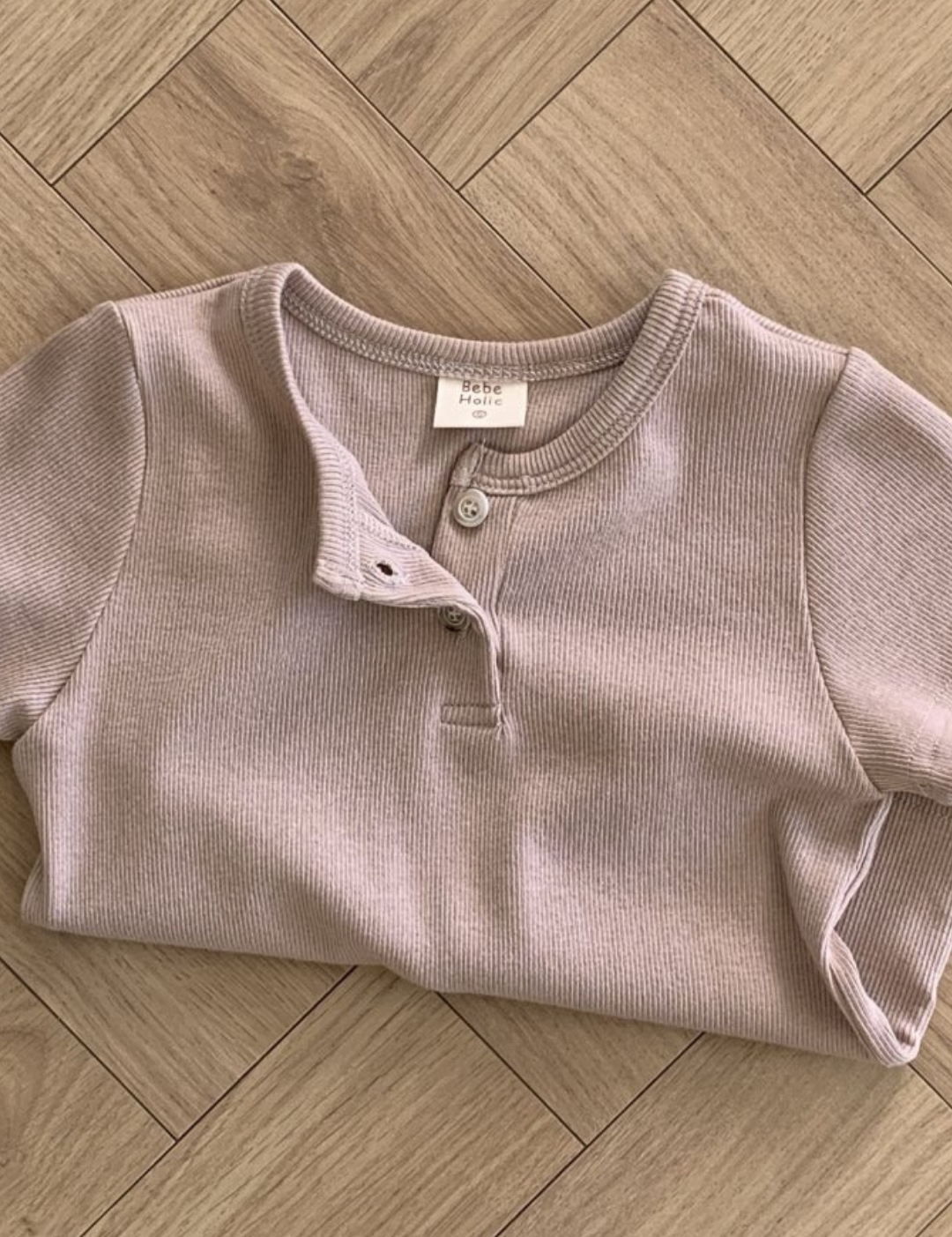 Ein weich aussehendes, kurzärmliges, geripptes Henley-Shirt in hellbeige Farbe liegt ordentlich gefaltet auf einem Holzparkettboden. Das Bebe Holic ST Tee besteht aus atmungsaktiver Baumwolle und hat einen geknöpften Ausschnitt mit einem offenen Knopf.