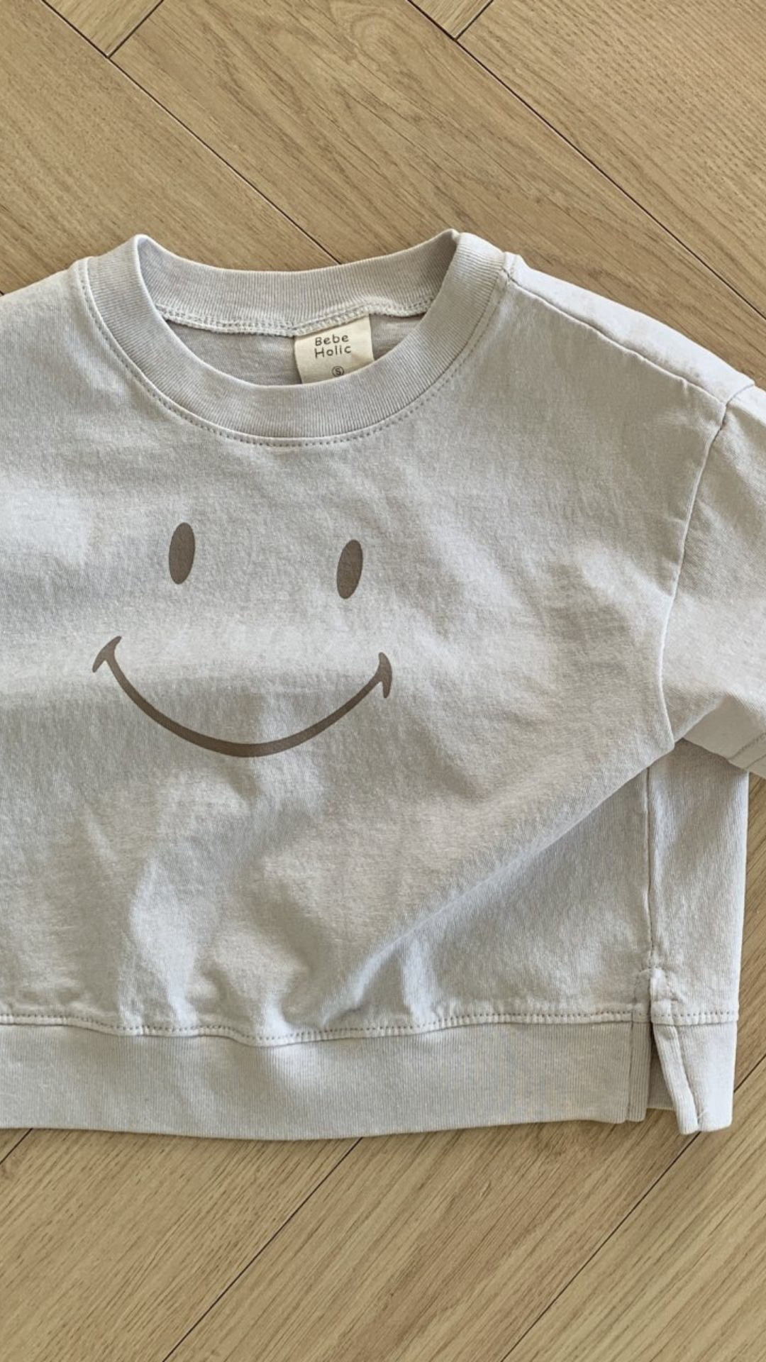 Ein hellgraues Sweatshirt aus weicher Baumwolle mit einem einfachen, großen Smiley-Design auf der Vorderseite wird ordentlich gefaltet und auf einen Holzboden gelegt. Auf dem Schild am Kragen steht „Smile Tee by Bebe Holic“.