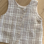 Eine ärmellose, karierte Weste mit Knopfleiste vorne liegt auf einem Holzboden. Das ärmellose Bebe Holic Summer Check Tee hat ein beige-braunes Karomuster und wird in der Mitte mit fünf Knöpfen geschlossen, was der Garderobe jedes Kindes einen Hauch modischen Flairs verleiht.