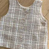 Eine ärmellose, karierte Weste mit Knopfleiste vorne liegt auf einem Holzboden. Das ärmellose Bebe Holic Summer Check Tee hat ein beige-braunes Karomuster und wird in der Mitte mit fünf Knöpfen geschlossen, was der Garderobe jedes Kindes einen Hauch modischen Flairs verleiht.