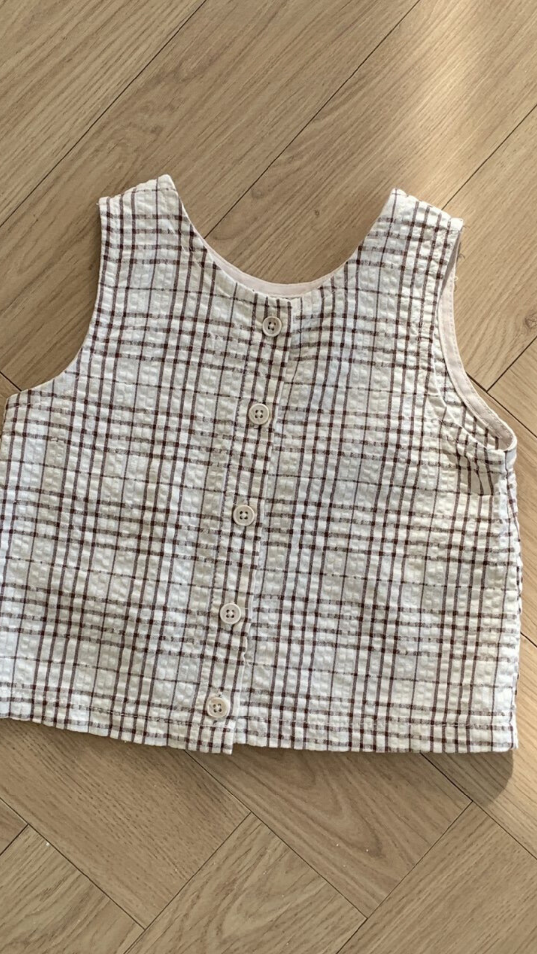 Eine ärmellose, karierte Weste mit Knopfleiste vorne liegt auf einem Holzboden. Das ärmellose Bebe Holic Summer Check Tee hat ein beige-braunes Karomuster und wird in der Mitte mit fünf Knöpfen geschlossen, was der Garderobe jedes Kindes einen Hauch modischen Flairs verleiht.