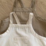 Zwei Bebeholic Twill Pocket Body Suits liegen flach auf einem Holzboden; einer ist taupe und der andere beige, beide mit Vordertaschen und Schulterriemen.