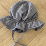 Ein grauer Crunch Bucket Hat aus der Bebe Holic-Kollektion mit breiter Krempe und Kinnriemen liegt auf einem Holzboden. Der Hut hat ein Gummiband um die Krone für einen gerafften Look, der seine natürliche Farbpalette perfekt ergänzt.