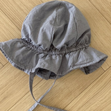 Ein grauer Crunch Bucket Hat aus der Bebe Holic-Kollektion mit breiter Krempe und Kinnriemen liegt auf einem Holzboden. Der Hut hat ein Gummiband um die Krone für einen gerafften Look, der seine natürliche Farbpalette perfekt ergänzt.