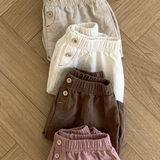 Ein Set aus fünf ordentlich gefalteten Paaren Bebe Holic Natural Pants in natürlichen Farbtönen, von oben nach unten: Hellbeige, Creme, Weiß, Dunkelbraun und Altrosa. Diese vielseitigen Must-have-Shorts haben eine elastische Taille und Knopfdetails. Sie liegen auf einem Holzboden.