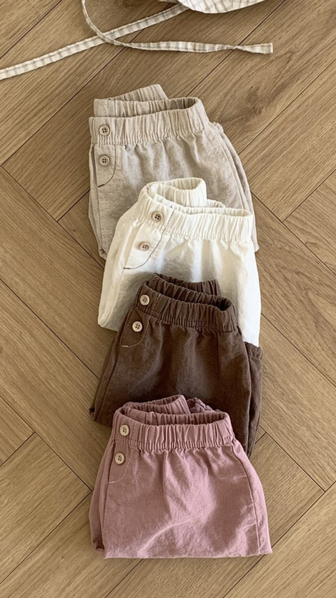 Ein Set aus fünf ordentlich gefalteten Paaren Bebe Holic Natural Pants in natürlichen Farbtönen, von oben nach unten: Hellbeige, Creme, Weiß, Dunkelbraun und Altrosa. Diese vielseitigen Must-have-Shorts haben eine elastische Taille und Knopfdetails. Sie liegen auf einem Holzboden.