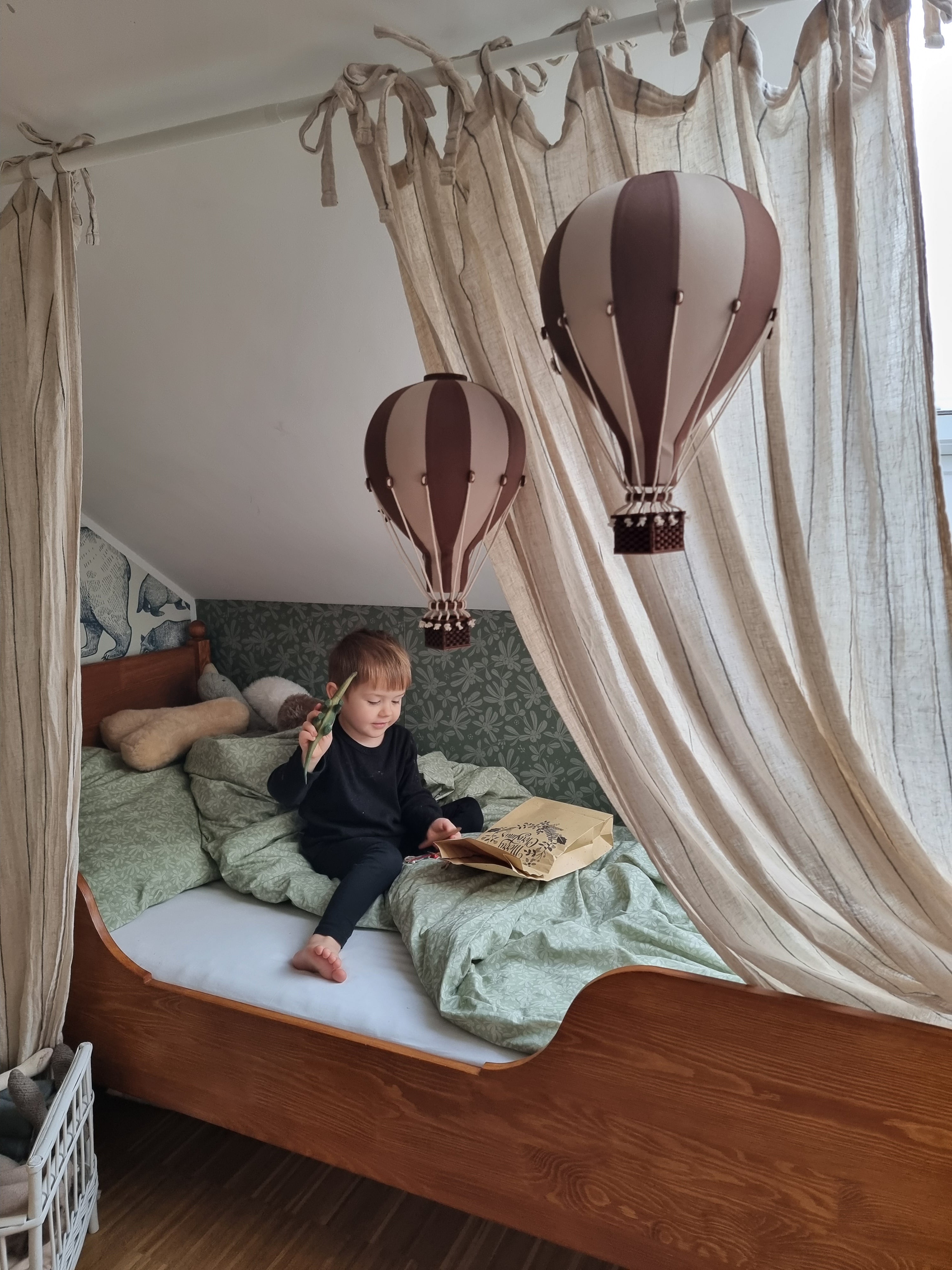 Ein kleines Kind sitzt auf einem Bett mit drapiertem Stoff darüber, hält ein grünes Blatt in der Hand und schaut in ein offenes Buch. Die kreative Raumgestaltung des Zimmers wird durch zwei dekorative SuperBalloons – Bunte Luftballons für magische Kinderzimmer-Momente, die über dem Bett hängen, hervorgehoben, und Stofftiere liegen neben dem Kind.