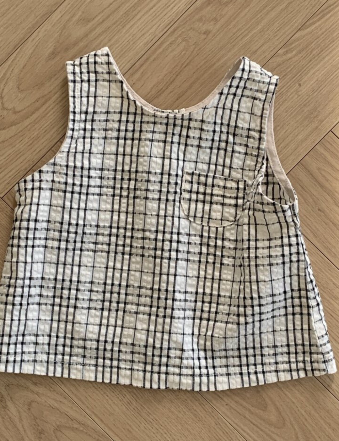 Ein Summer Check Sleeveless Tee von Bebe Holic mit weißem Hintergrund und schwarzem Gittermuster liegt flach auf einem Holzboden und verströmt modisches Flair. Das Oberteil verfügt über eine kleine Vordertasche auf der linken Seite, die jedem Outfit einen Hauch von Eleganz verleiht.