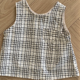 Ein Summer Check Sleeveless Tee von Bebe Holic mit weißem Hintergrund und schwarzem Gittermuster liegt flach auf einem Holzboden und verströmt modisches Flair. Das Oberteil verfügt über eine kleine Vordertasche auf der linken Seite, die jedem Outfit einen Hauch von Eleganz verleiht.