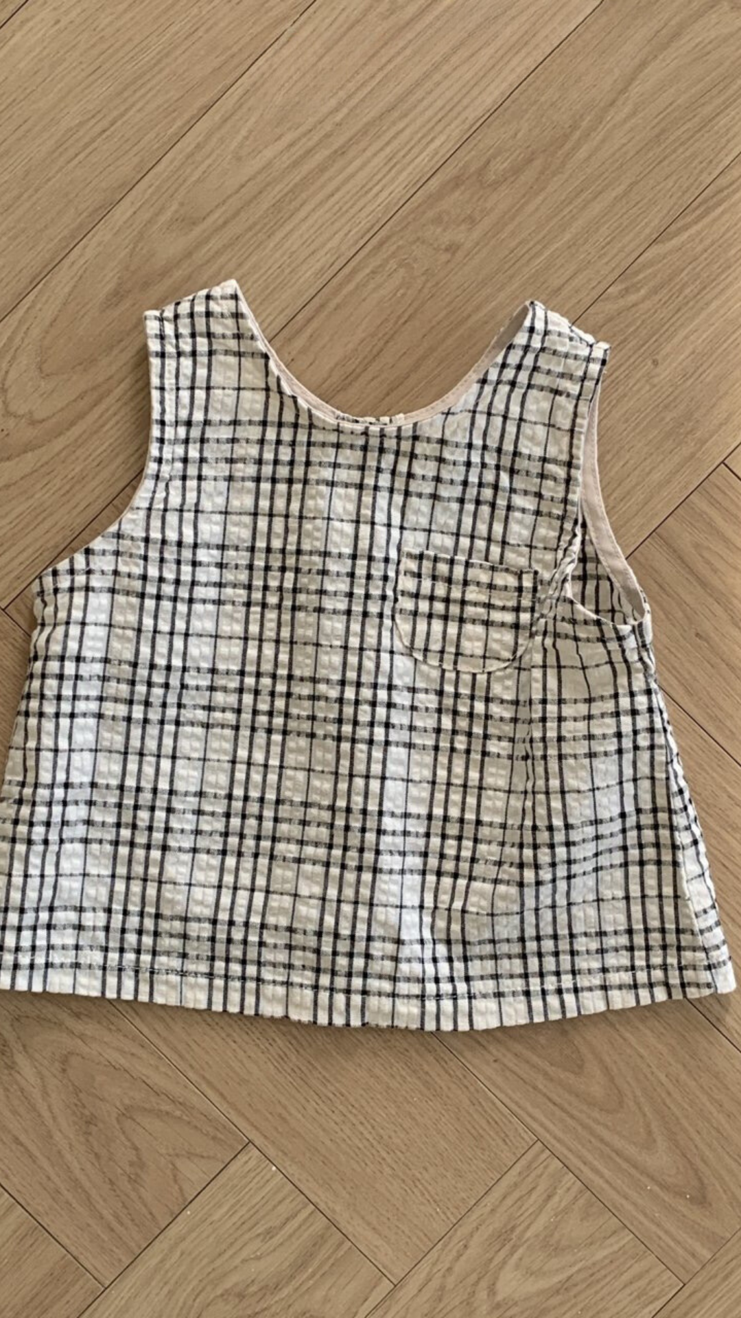 Ein Summer Check Sleeveless Tee von Bebe Holic mit weißem Hintergrund und schwarzem Gittermuster liegt flach auf einem Holzboden und verströmt modisches Flair. Das Oberteil verfügt über eine kleine Vordertasche auf der linken Seite, die jedem Outfit einen Hauch von Eleganz verleiht.