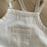 Eine beige, ärmellose Schürze aus robustem Stoff liegt flach auf einer Holzoberfläche. Die Schürze, die an den Bebe Holic Twill Pocket Body Suit erinnert, verfügt über eine kleine, rechteckige Tasche auf der Brust und hat zwei verstellbare Träger. Die natürliche Beleuchtung hebt die Textur des Stoffes und die Holzmaserung hervor.