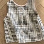 Ein ärmelloses, kragenloses, schwarz-weiß kariertes Hemd mit kleinen Knöpfen liegt flach auf einem Holzboden mit einem Fischgrätenmuster. Das Design dieses ärmellosen Bebe Holic Summer Check Tee zeigt ein einfaches Gittermuster aus schwarzen Linien auf weißem Hintergrund und verleiht jedem Outfit modisches Flair.