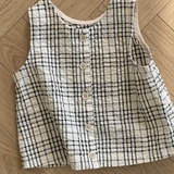 Ein ärmelloses, kragenloses, schwarz-weiß kariertes Hemd mit kleinen Knöpfen liegt flach auf einem Holzboden mit einem Fischgrätenmuster. Das Design dieses ärmellosen Bebe Holic Summer Check Tee zeigt ein einfaches Gittermuster aus schwarzen Linien auf weißem Hintergrund und verleiht jedem Outfit modisches Flair.