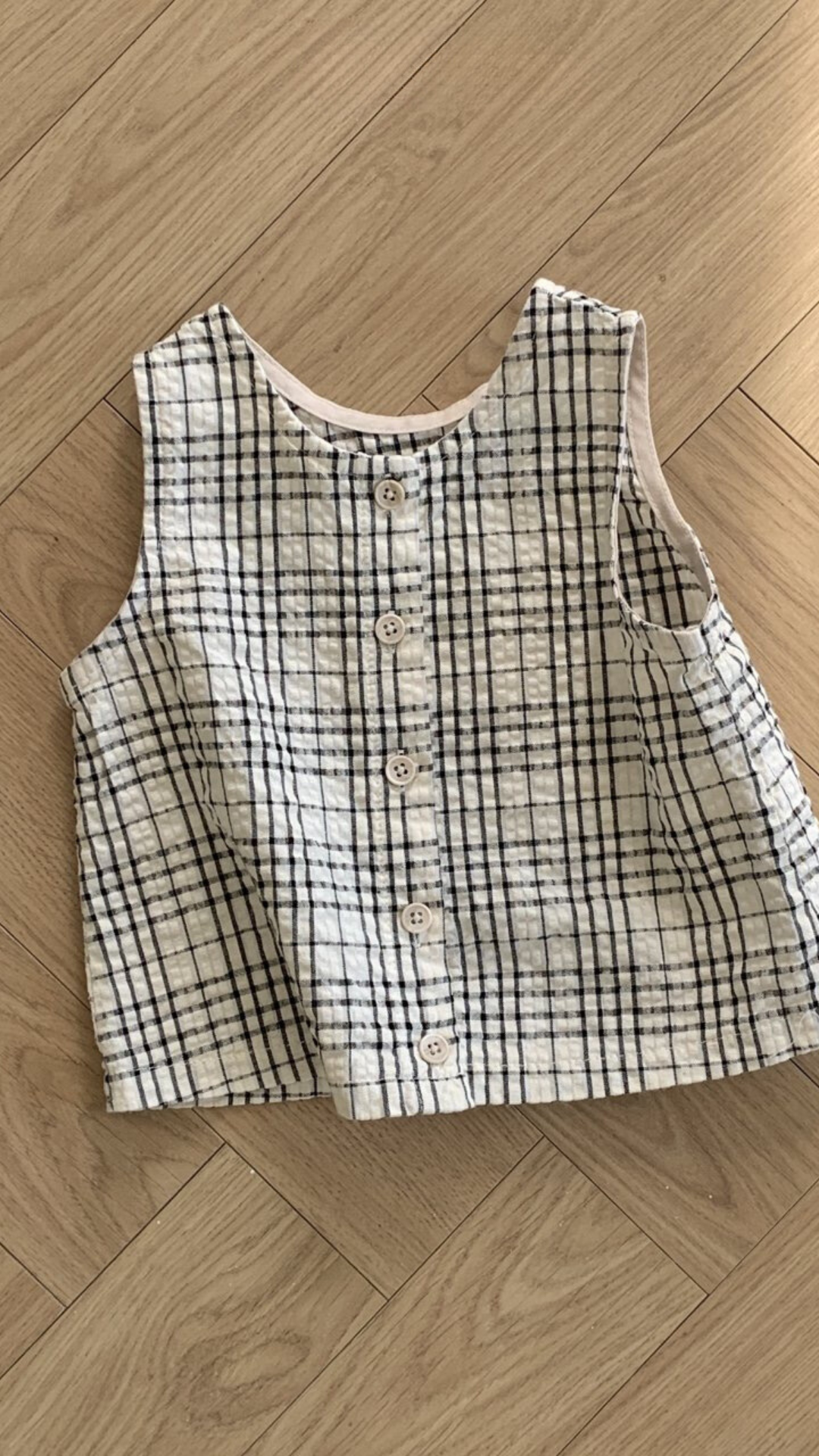 Ein ärmelloses, kragenloses, schwarz-weiß kariertes Hemd mit kleinen Knöpfen liegt flach auf einem Holzboden mit einem Fischgrätenmuster. Das Design dieses ärmellosen Bebe Holic Summer Check Tee zeigt ein einfaches Gittermuster aus schwarzen Linien auf weißem Hintergrund und verleiht jedem Outfit modisches Flair.