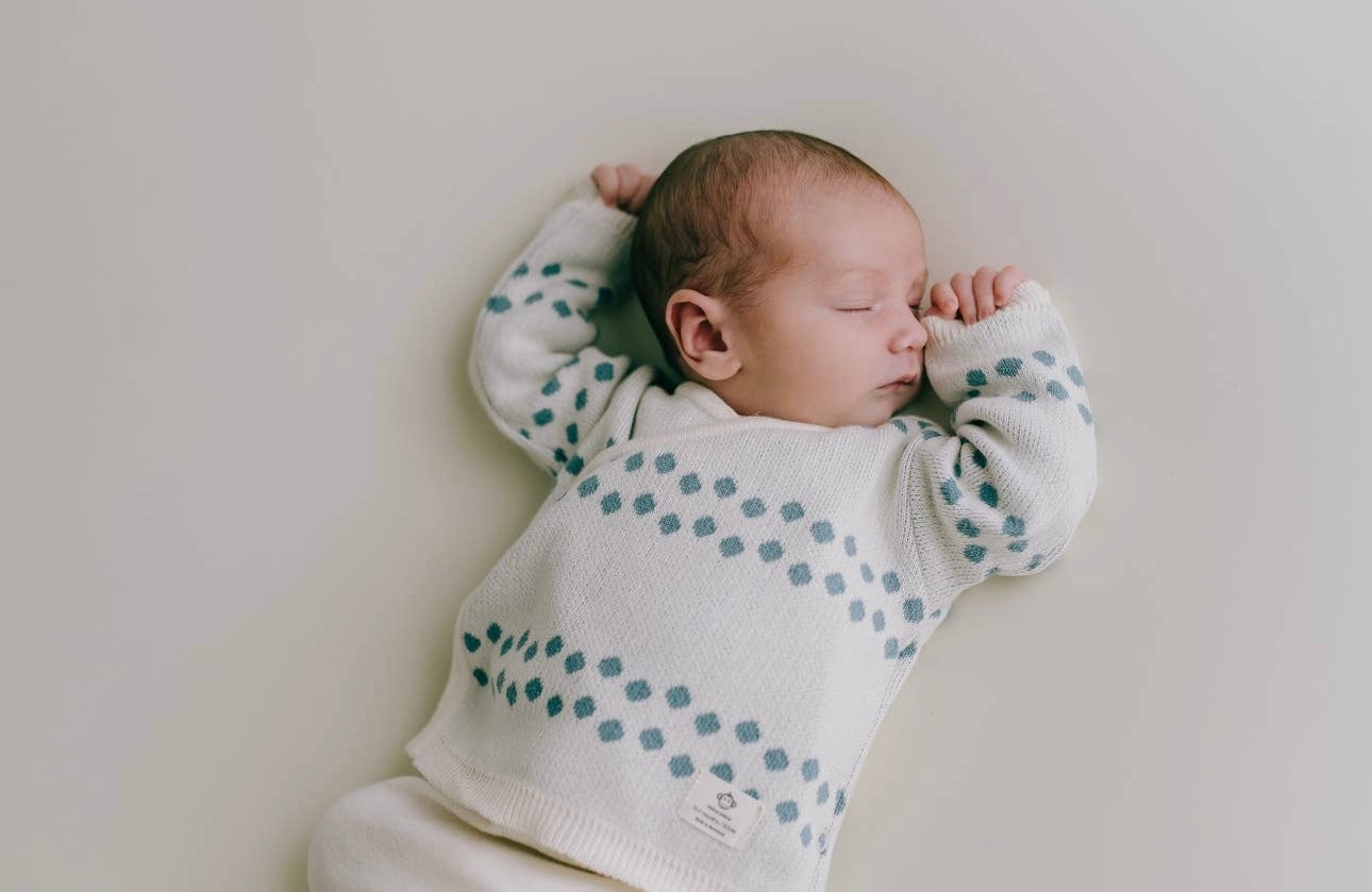 Ein Neugeborenes schläft friedlich, gekleidet in einen weißen Pullover mit blauen Punkten aus 100 % Bio-Baumwolle, der vor einem weichen, hellen Hintergrund liegt. Außerdem trägt das Baby Pack Dots Jeans für Neugeborene von micu micu.