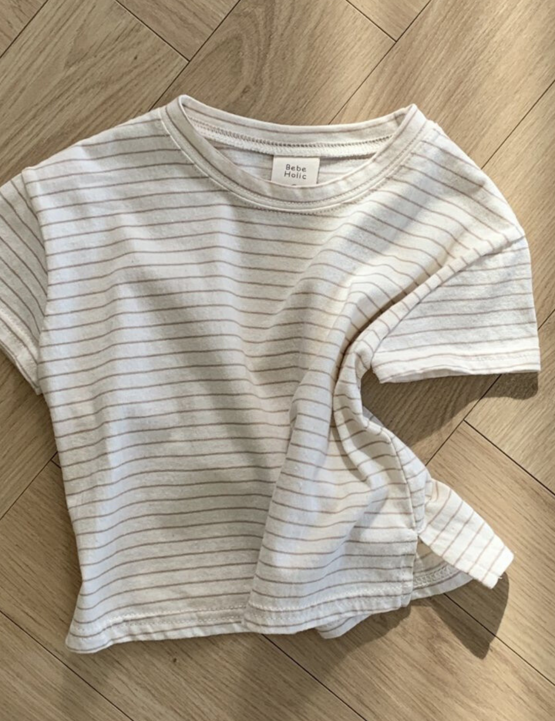 Ein weißes kurzärmliges Basic ST Tee mit dünnen schwarzen Querstreifen von Bebe Holic liegt ordentlich auf einem Holzboden mit Fischgrätenmuster und zeigt seinen klassischen Stil. Der Stoff des T-Shirts ist leicht zerknittert.