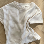 Ein weißes kurzärmliges Basic ST Tee mit dünnen schwarzen Querstreifen von Bebe Holic liegt ordentlich auf einem Holzboden mit Fischgrätenmuster und zeigt seinen klassischen Stil. Der Stoff des T-Shirts ist leicht zerknittert.