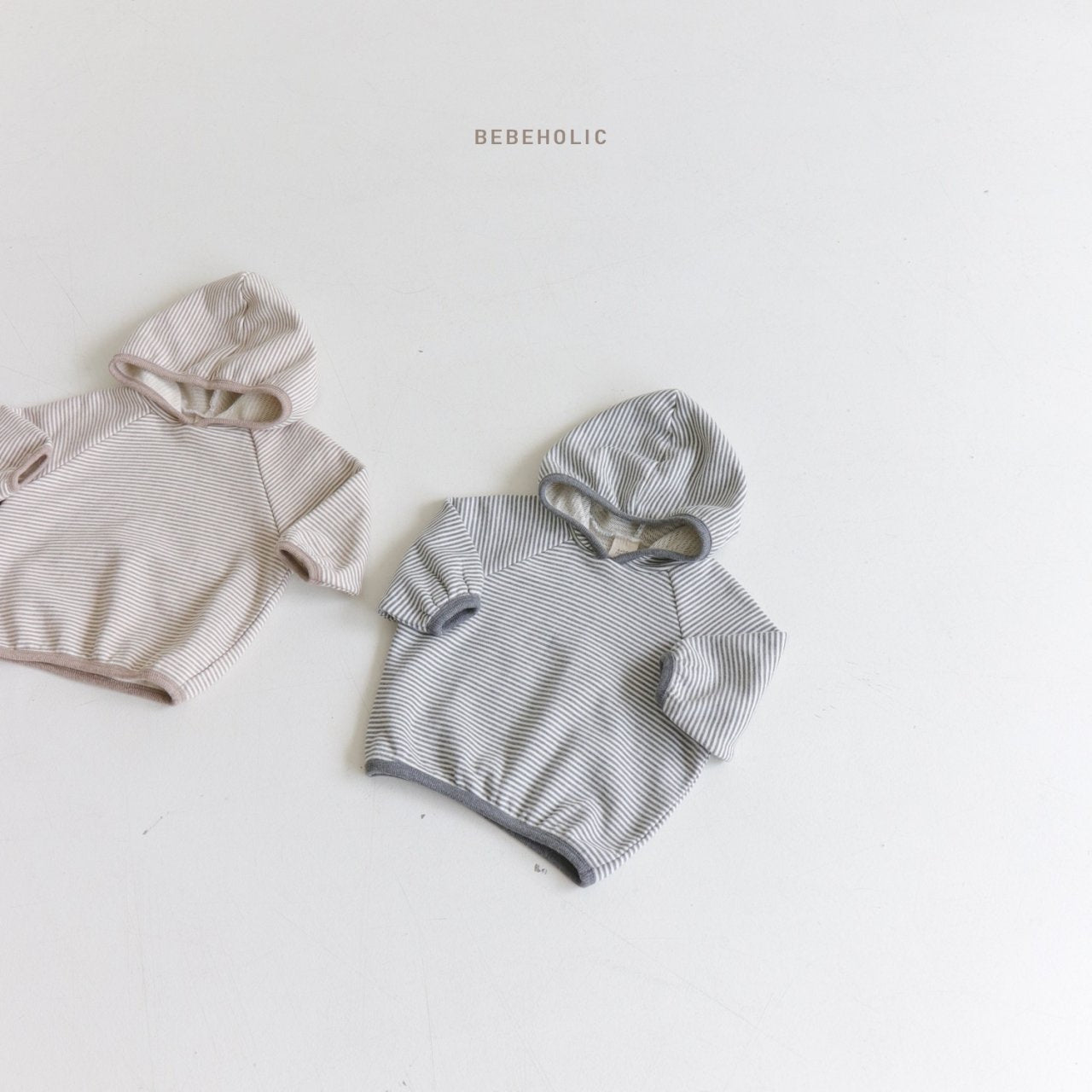 Zwei Kleinkinderhoodies in Hellbraun und Grau liegen nebeneinander auf einem weißen Untergrund. Beide gemütlichen Hoodies sind quergestreift und haben Kapuzen. Über dem niedlichen Macaroon Hoody für Babys steht „Bebe Holic“.