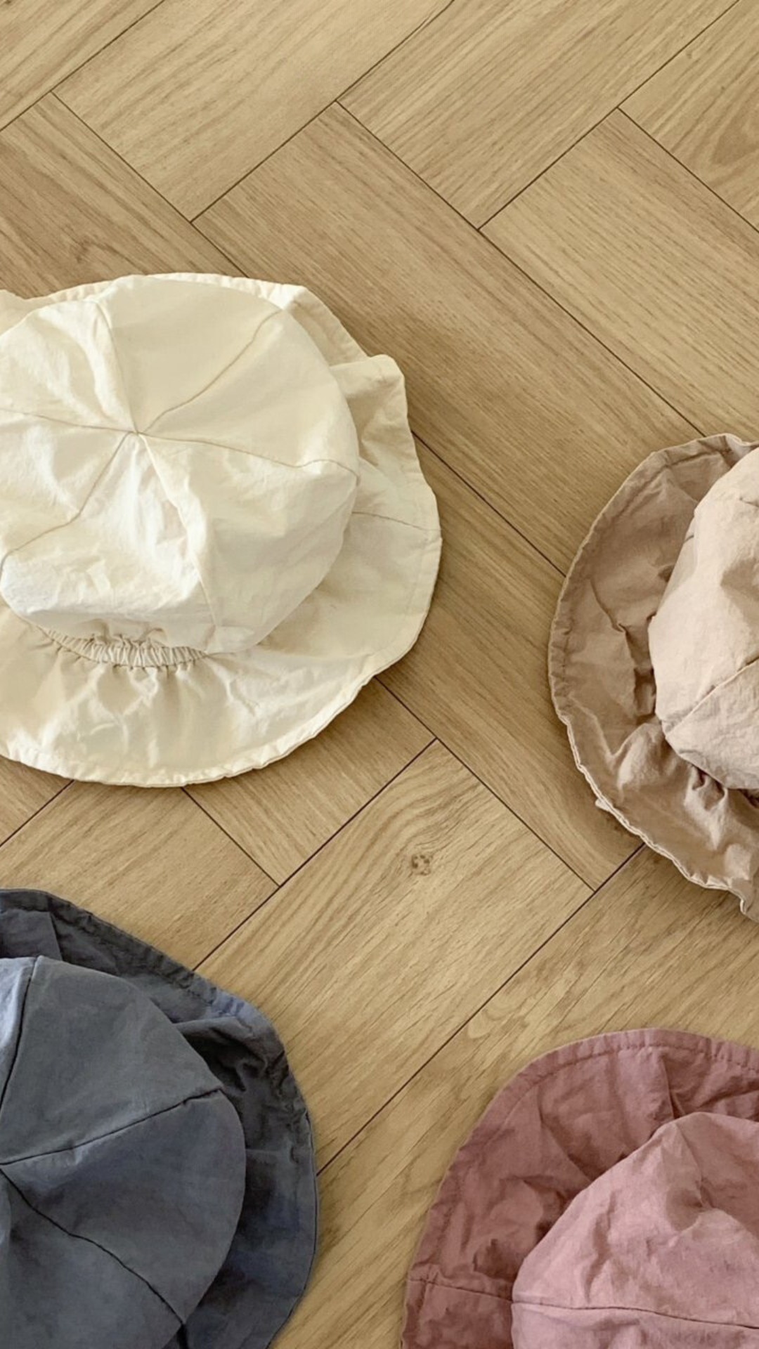 Vier zerknitterte Crunch Bucket Hats von Bebe Holic in einer natürlichen Farbpalette aus Weiß, Beige, Blau und Kastanienbraun sind auf einem hölzernen Boden mit Fischgrätenmuster ausgebreitet. Die Hüte scheinen aus einem leichten, lässigen Stoff gefertigt zu sein.