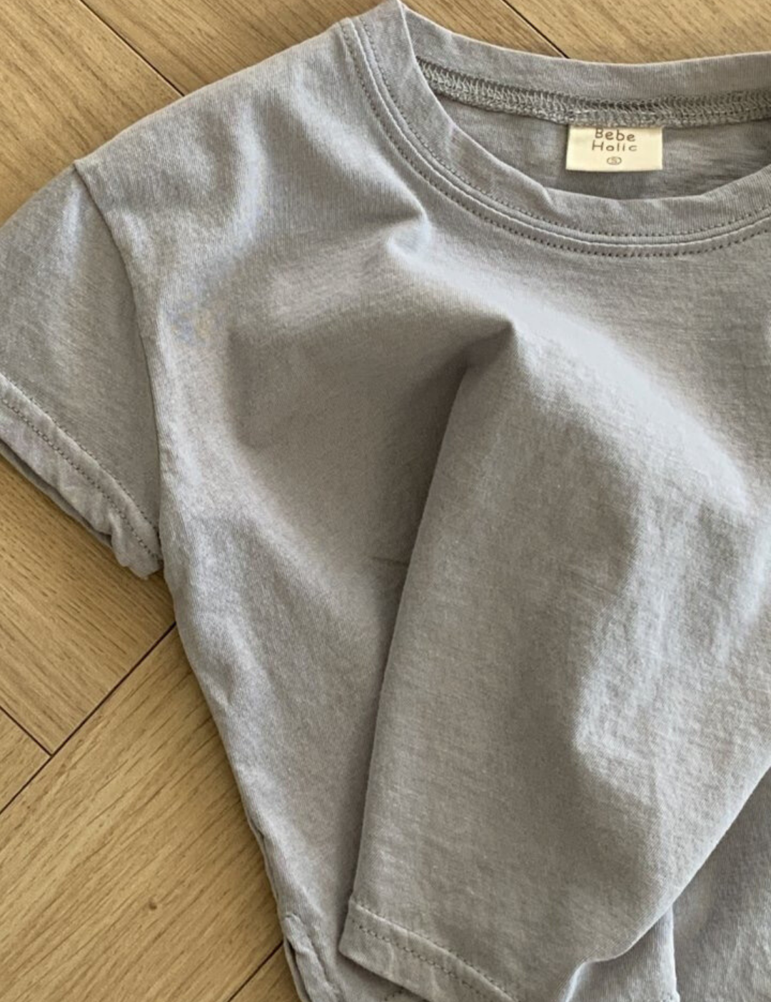 Ein hellgraues Basic Tee von Bebe Holic aus hochwertiger Baumwolle liegt zerknüllt auf einem Holzboden. Das Shirt hat kurze Ärmel und einen runden Ausschnitt, in natürlichen Tönen. Im Krageninneren ist ein kleines weißes Etikett sichtbar.