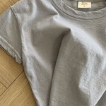 Ein hellgraues Basic Tee von Bebe Holic aus hochwertiger Baumwolle liegt zerknüllt auf einem Holzboden. Das Shirt hat kurze Ärmel und einen runden Ausschnitt, in natürlichen Tönen. Im Krageninneren ist ein kleines weißes Etikett sichtbar.