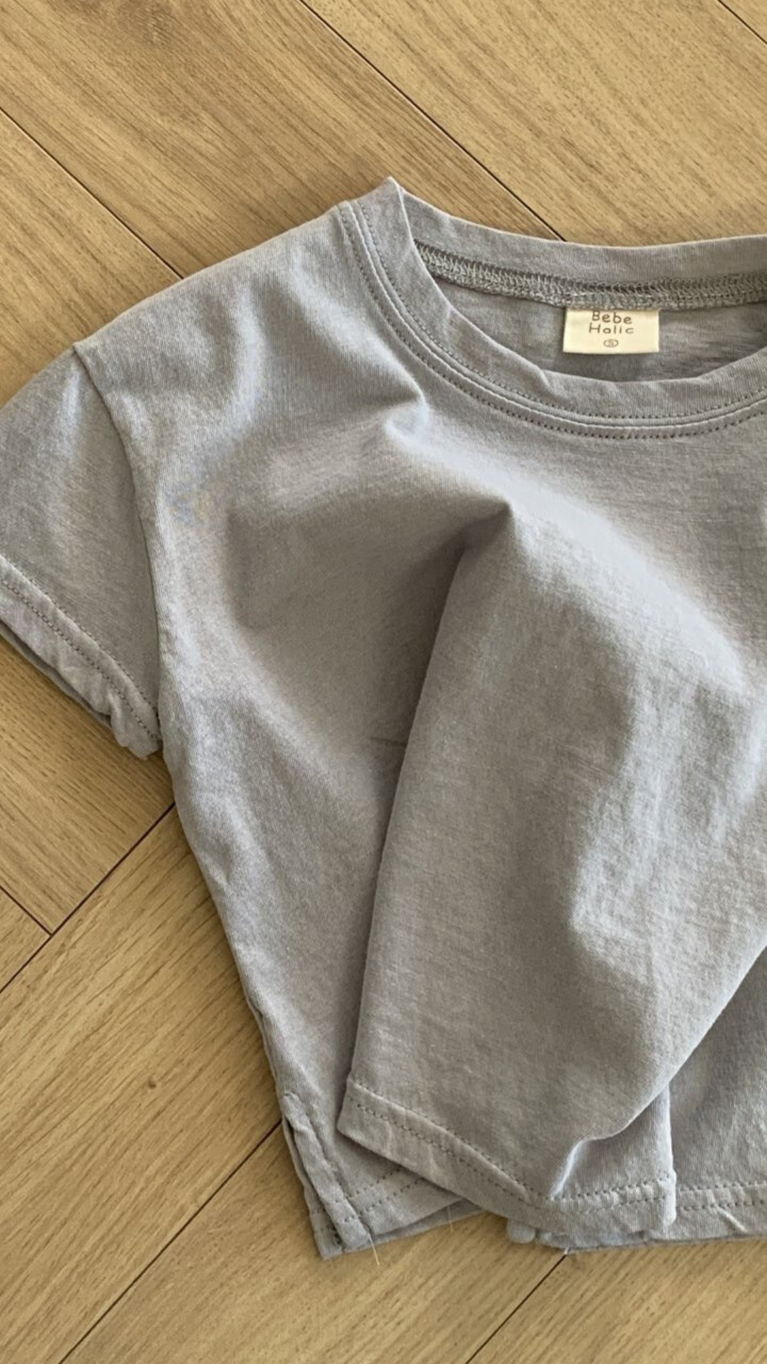 Ein hellgraues Basic Tee von Bebe Holic aus hochwertiger Baumwolle liegt zerknüllt auf einem Holzboden. Das Shirt hat kurze Ärmel und einen runden Ausschnitt, in natürlichen Tönen. Im Krageninneren ist ein kleines weißes Etikett sichtbar.