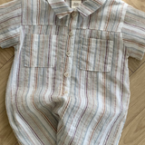 Ein kurzärmeliger Aurora-Bodysuit von Bebe Holic liegt auf einem Holzboden. Der Strampler, der an gestreifte Hemden erinnert, hat einen Kragen, zwei Brusttaschen und Knöpfe auf der Vorderseite. Er besticht durch ein Streifenmuster in verschiedenen Blau-, Rot- und Beigetönen.