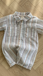 Ein kurzärmeliger Aurora-Bodysuit von Bebe Holic liegt auf einem Holzboden. Der Strampler, der an gestreifte Hemden erinnert, hat einen Kragen, zwei Brusttaschen und Knöpfe auf der Vorderseite. Er besticht durch ein Streifenmuster in verschiedenen Blau-, Rot- und Beigetönen.