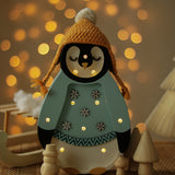 Festliche Little Lights – Baby Penguin Mini Christmas Edition | Blau mit leuchtenden Lichtern trägt eine senfgelbe Strickmütze und einen blauen Pullover mit Schneeflocken. Er steht auf einer Holzoberfläche mit verschwommenen Bokeh-Lichtern im Hintergrund und schafft eine warme und gemütliche Atmosphäre, perfekt als Kinderzimmer-Dekoration.