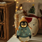 Eine gemütliche Weihnachtsszene mit einer dekorativen Pinguinfigur, die eine gelbe Mütze und einen blauen Pullover mit Schneeflockenmotiven trägt. Die Little Lights – Baby Penguin Mini Christmas Edition | Blue strahlt ein warmes Licht aus, perfekt für die Kinderzimmer-Dekoration, und im Hintergrund befinden sich festliche Kissen auf einem Korbstuhl mit einem Weihnachtsbaummotiv.