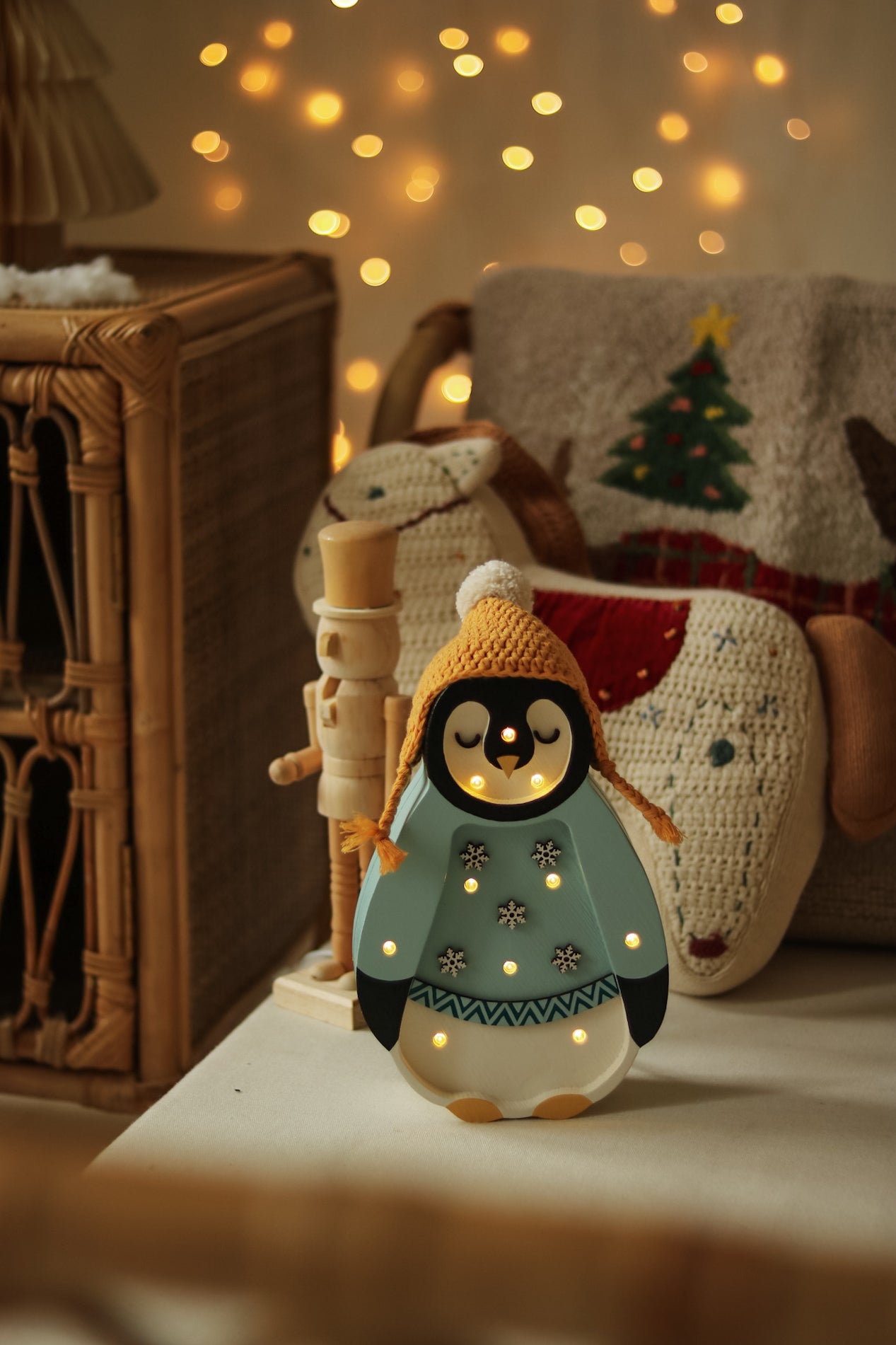 Eine gemütliche Weihnachtsszene mit einer dekorativen Pinguinfigur, die eine gelbe Mütze und einen blauen Pullover mit Schneeflockenmotiven trägt. Die Little Lights – Baby Penguin Mini Christmas Edition | Blue strahlt ein warmes Licht aus, perfekt für die Kinderzimmer-Dekoration, und im Hintergrund befinden sich festliche Kissen auf einem Korbstuhl mit einem Weihnachtsbaummotiv.