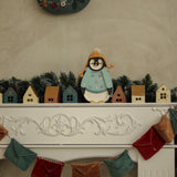 Auf einem festlichen Kaminsims steht ein Little Lights - Baby Penguin Mini Christmas Edition | Blue von Little Lights mit Wintermantel und Mütze zwischen Miniatur-Holzhäusern und grünen Tannenzweigen. Darüber hängt ein Weihnachtskranz. Darunter hängen bunte Stoffwimpel an der Vorderseite des Kaminsimses und bilden die perfekte Kinderzimmer-Dekoration.