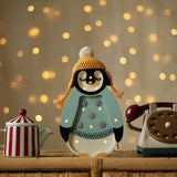 Eine festliche Szene zeigt eine süße Pinguinfigur, die eine gelbe Strickmütze mit Bommel und einen hellblauen Mantel mit Schneeflocken trägt. Der Pinguin, der gleichzeitig ein entzückender Little Lights - Baby Penguin Mini Christmas Edition | Blue von Little Lights ist, ist von einer rot-weiß gestreiften Teekanne und einem Vintage-Wählscheibentelefon auf einer Korboberfläche umgeben, mit funkelnden Lichtern im Hintergrund.