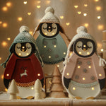 Drei entzückende Pinguinfiguren sind in gemütliche Winteroutfits gekleidet, komplett mit Strickmützen und gestickten Details. Erleuchtet von winzigen Lichtern dient diese bezaubernde Little Lights - Baby Penguin Mini Christmas Edition | Blue von Little Lights auch als Nachtlicht, umgeben von festlichem, leuchtendem Dekor vor einem verschwommenen Hintergrund, gesprenkelt mit warmen, herzförmigen Lichtern.