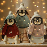 Drei entzückende Pinguinfiguren sind in gemütliche Winteroutfits gekleidet, komplett mit Strickmützen und gestickten Details. Erleuchtet von winzigen Lichtern dient diese bezaubernde Little Lights - Baby Penguin Mini Christmas Edition | Blue von Little Lights auch als Nachtlicht, umgeben von festlichem, leuchtendem Dekor vor einem verschwommenen Hintergrund, gesprenkelt mit warmen, herzförmigen Lichtern.