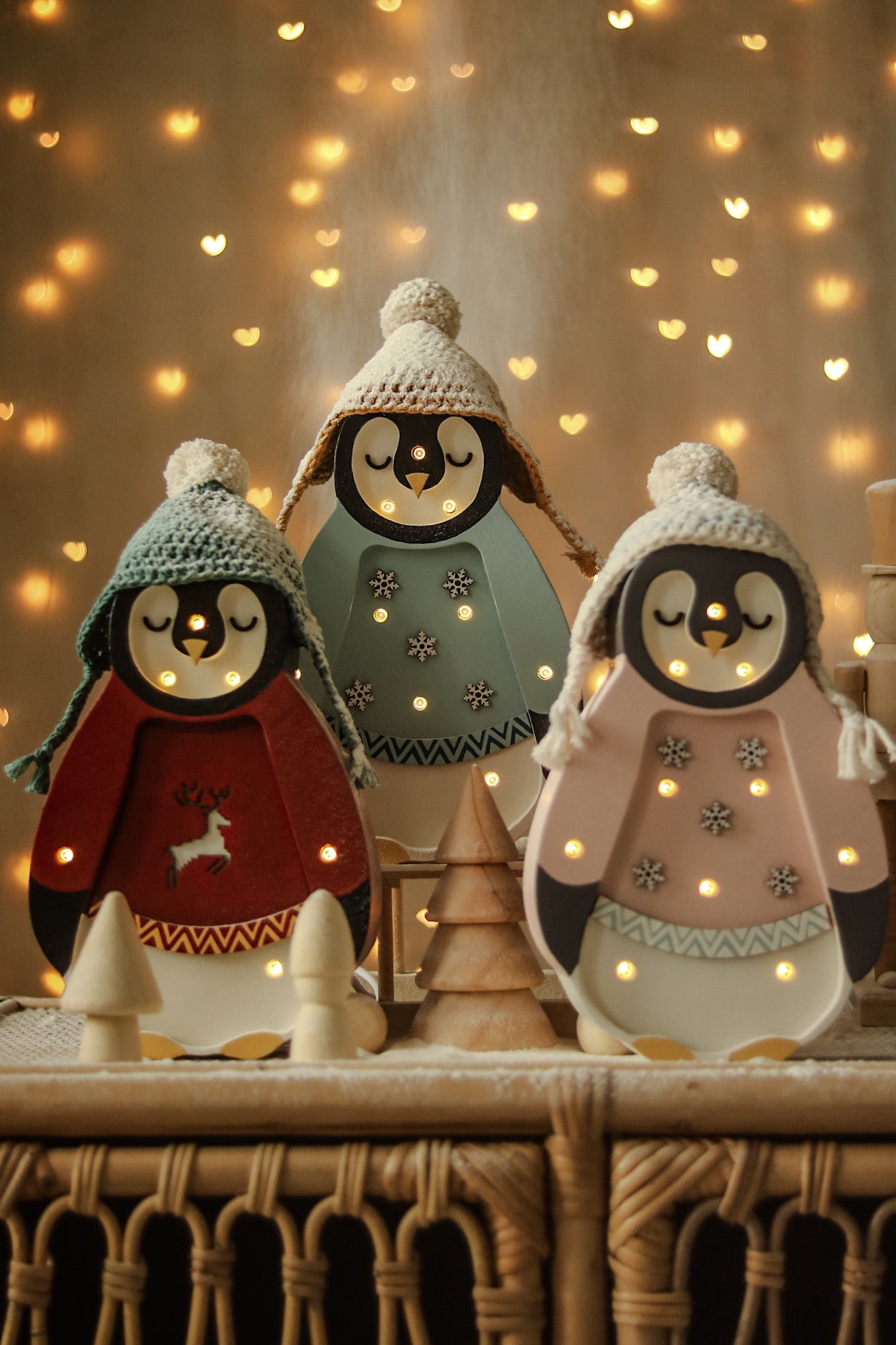 Drei entzückende Pinguinfiguren sind in gemütliche Winteroutfits gekleidet, komplett mit Strickmützen und gestickten Details. Erleuchtet von winzigen Lichtern dient diese bezaubernde Little Lights - Baby Penguin Mini Christmas Edition | Blue von Little Lights auch als Nachtlicht, umgeben von festlichem, leuchtendem Dekor vor einem verschwommenen Hintergrund, gesprenkelt mit warmen, herzförmigen Lichtern.