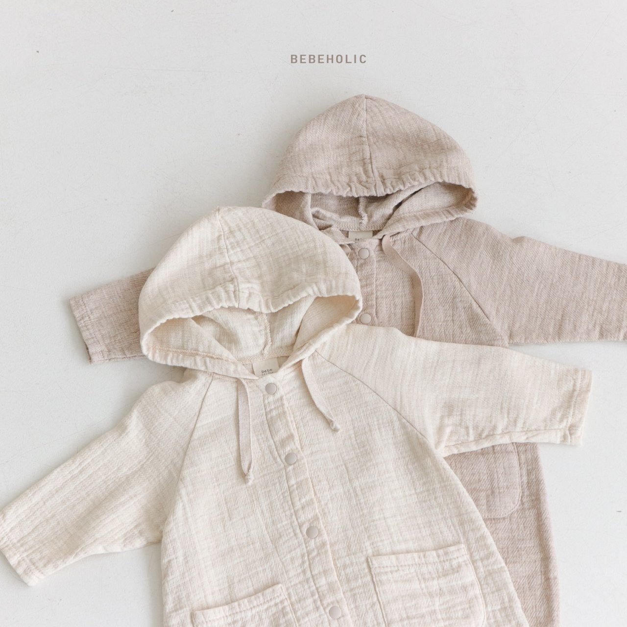 Zwei Bebe Holic Hoody Bodysuits liegen flach auf einer hellen Fläche. Der obere Onesie ist hellbeige, der untere etwas dunkleres Beige. Beide haben Knöpfe vorne und Taschen. Über den Onesies ist der Markenname „Bebe Holic“ aufgedruckt.