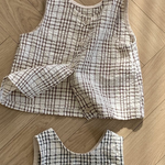 Zwei ärmellose, karierte Westen aus gestepptem Stoff werden auf einem Holzboden ausgestellt. Die obere Weste aus der Summer Check Sleeveless Tee-Kollektion von Bebe Holic hat ein braun-weißes Muster mit Knöpfen auf der Vorderseite, während die untere Weste ein schwarz-weißes Muster ohne sichtbare Knöpfe aufweist, was modisches Flair verleiht.