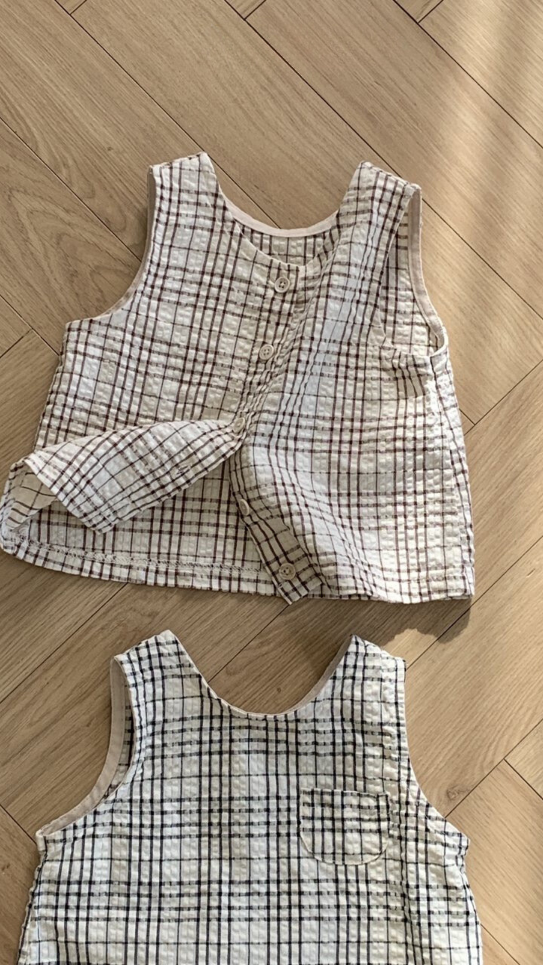 Zwei ärmellose, karierte Westen aus gestepptem Stoff werden auf einem Holzboden ausgestellt. Die obere Weste aus der Summer Check Sleeveless Tee-Kollektion von Bebe Holic hat ein braun-weißes Muster mit Knöpfen auf der Vorderseite, während die untere Weste ein schwarz-weißes Muster ohne sichtbare Knöpfe aufweist, was modisches Flair verleiht.