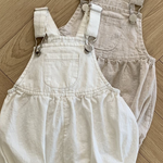 Zwei Bebe Holic Leinen-Latzhosen mit Schnallen, eine in Off-White und die andere in Beige, liegen auf einem hellen Holzboden. Jeder Strampler aus atmungsaktiver Baumwolle hat verstellbare Träger mit Metallschnallen und eine Vordertasche mit gerafftem Stoff um die Taille und Beinöffnungen.