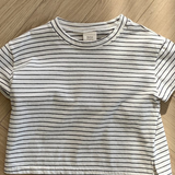 Ein kurzärmliges, weißes Crop-Top mit schwarzen Querstreifen liegt flach auf einem Holzboden. Dieses Basic ST Tee von Bebe Holic hat einen runden Ausschnitt und ein Etikett im Kragen. Der Holzboden mit seinem hellen, natürlichen Finish und Fischgrätenmuster unterstreicht seinen klassischen Stil und bequemen Tragekomfort.