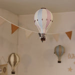 Ein mit vielseitiger Dekoration dekorierter Raum mit hängenden Ornamenten in Form von Heißluftballons. Im Vordergrund ist ein blau-rosa gestreifter dekorativer Ballon – Beige, Mint, Grau und Lila | Einzigartiges Heißluftballon-Ornament für Ihr Zuhause von Superballoon zu sehen, im Hintergrund sind weitere dekorative Ballons in gedeckten Farben zu sehen. Ein dreieckiges Wimpelbanner hängt anmutig quer durch den Raum.